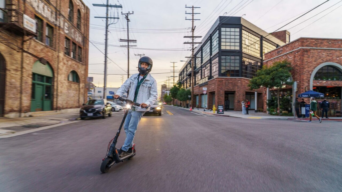 Uomo con casco integrale nero su un monopattino elettrico con il faro acceso in una strada di città al tramonto