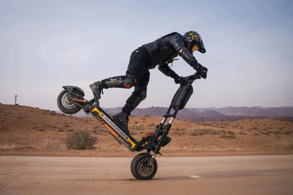 Pilota con casco e protezioni solleva la ruota posteriore di un monopattino elettrico KuKirin nero e arancio nel deserto