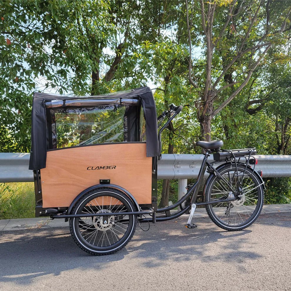 Cargo bike a tre ruote con cassone in legno e capottina nera con finestre, ferma vicino a un guardrail tra alberi verdi