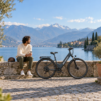 Bicicletta elettrica OneSport OT12 Pro su un molo acciottolato in riva a un lago, con un uomo seduto sul muretto accanto e montagne sullo sfondo