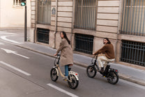 Due persone in bicicletta su una strada cittadina, ciascuna su una bicicletta pieghevole, una beige e una nera