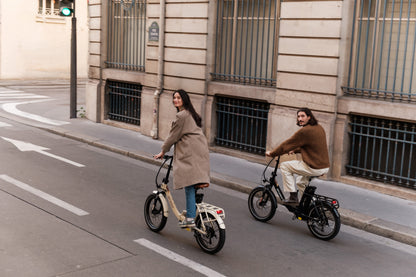 Due persone in bicicletta su una strada cittadina, ciascuna su una bicicletta pieghevole, una beige e una nera