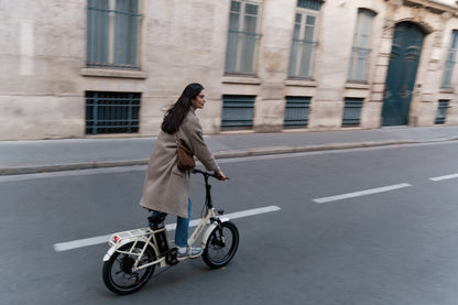 OneSport OT16 Pro: donna con cappotto in bicicletta su una strada davanti a una facciata in pietra, vista laterale