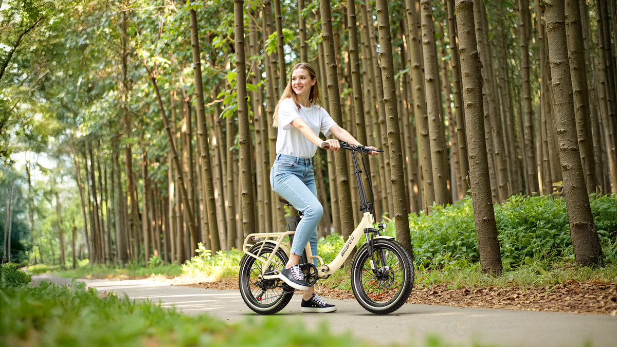 Donna sorridente accanto a una OneSport OT16 Pro color crema su un sentiero nel bosco tra alti tronchi d'albero