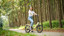 Donna sorridente accanto a una OneSport OT16 Pro color crema su un sentiero nel bosco tra alti tronchi d'albero