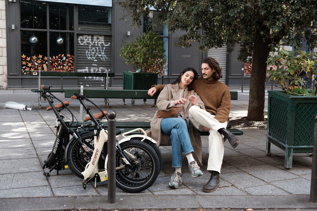 Una coppia seduta su una panchina accanto a due biciclette OneSport pieghevoli, una beige e una nera