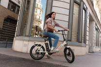 Donna in sella a una OneSport OT16 Pro, bicicletta elettrica bianca, vista di lato su una strada cittadina accanto a un edificio in pietra