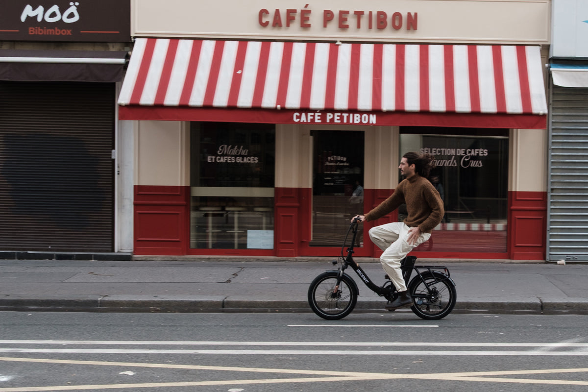 Uomo in sella a una OneSport OT16 Pro, bicicletta nera, in vista laterale davanti a un caffè con tenda a righe rosse e bianche
