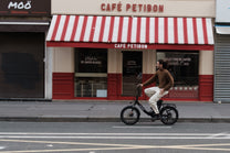 Uomo in sella a una OneSport OT16 Pro, bicicletta nera, in vista laterale davanti a un caffè con tenda a righe rosse e bianche