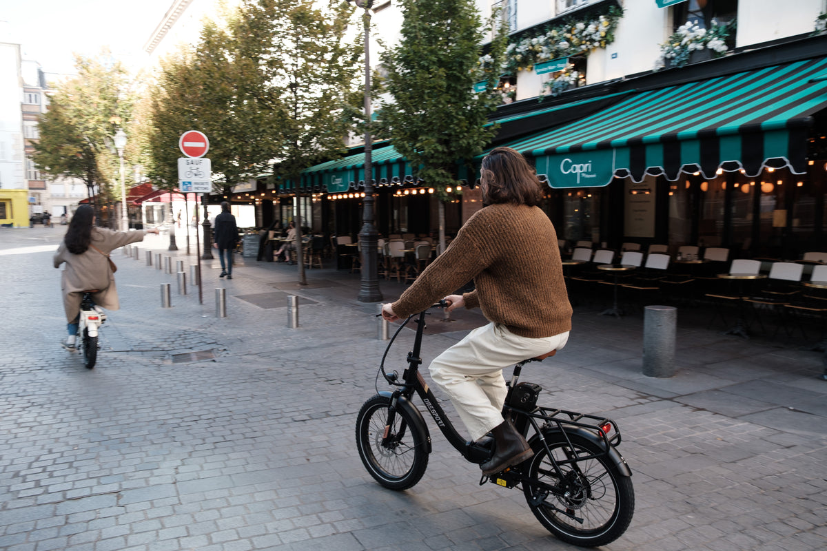 Persona in sella a una OneSport OT16 Pro, bicicletta nera, su una strada acciottolata davanti a un caffè con tenda a righe verdi