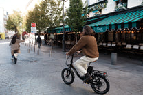 Persona in sella a una OneSport OT16 Pro, bicicletta nera, su una strada acciottolata davanti a un caffè con tenda a righe verdi