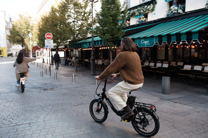 Persona in sella a una OneSport OT16 Pro, bicicletta nera, su una strada acciottolata davanti a un caffè con tenda a righe verdi