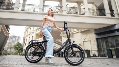 OneSport OT16 Pro: bicicletta nera in vista laterale con una donna in piedi accanto a essa su una piazza lastricata
