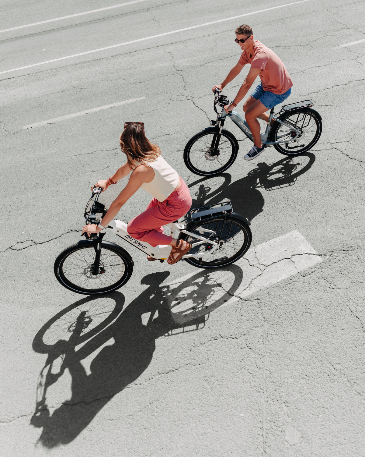 OneSport WT05 vista dall'alto, donna su bicicletta bianca e uomo su bicicletta grigia pedalano sull'asfalto con le ombre