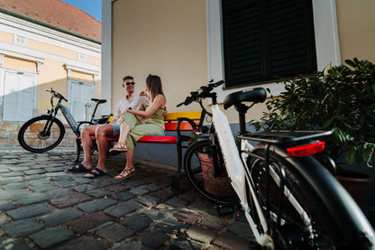 OneSport WT05 bianca in primo piano, una coppia seduta su una panchina colorata e un'altra bicicletta dietro su una strada acciottolata