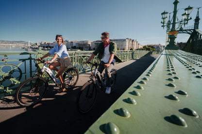 Due persone in bicicletta su un ponte verde al sole con edifici e acqua sullo sfondo