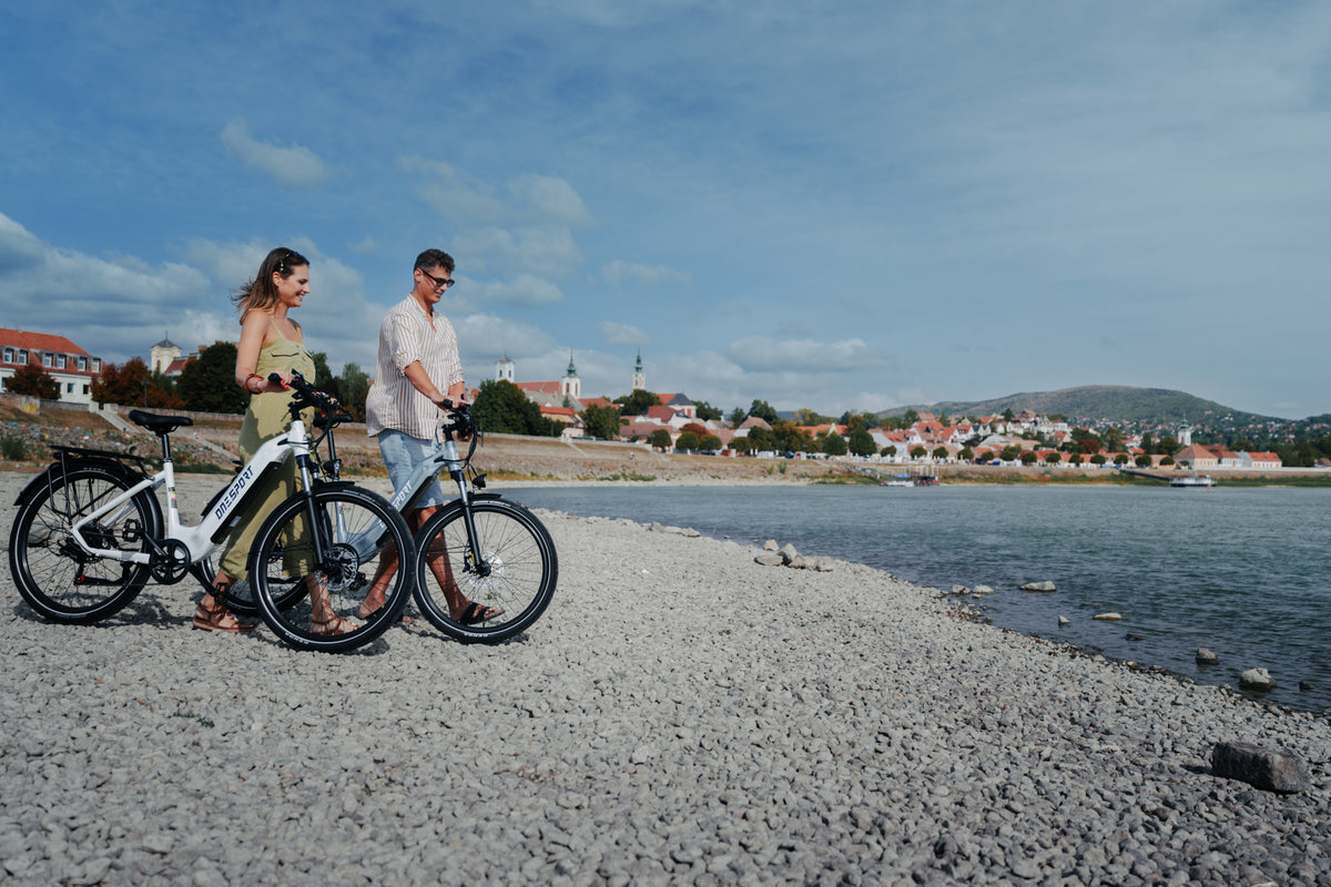 Due persone conducono una OneSport WT05 ciascuna lungo una spiaggia di pietre con città e collina sullo sfondo