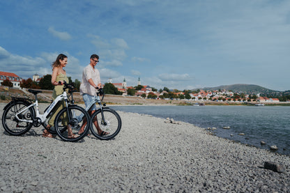 Due persone conducono una OneSport WT05 ciascuna lungo una spiaggia di pietre con città e collina sullo sfondo