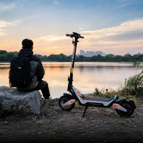 Wheely - J18 sul cavalletto laterale vicino a un lago al tramonto, con una persona con zaino seduta su una roccia accanto