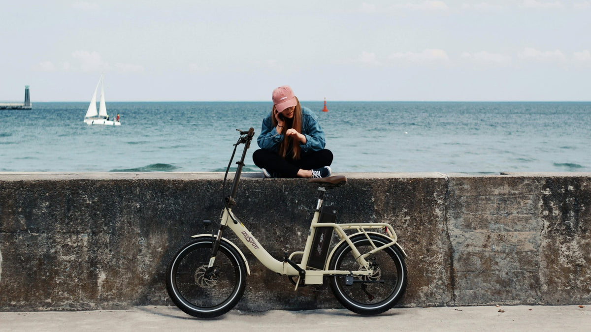 Uomo in piedi con la bici elettrica pieghevole OneSport OT16-2 in colore beige in riva al mare