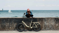 Uomo in piedi con la bici elettrica pieghevole OneSport OT16-2 in colore beige in riva al mare