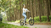 Donna alla guida della bici elettrica pieghevole OneSport OT16-2 in colore beige nel bosco