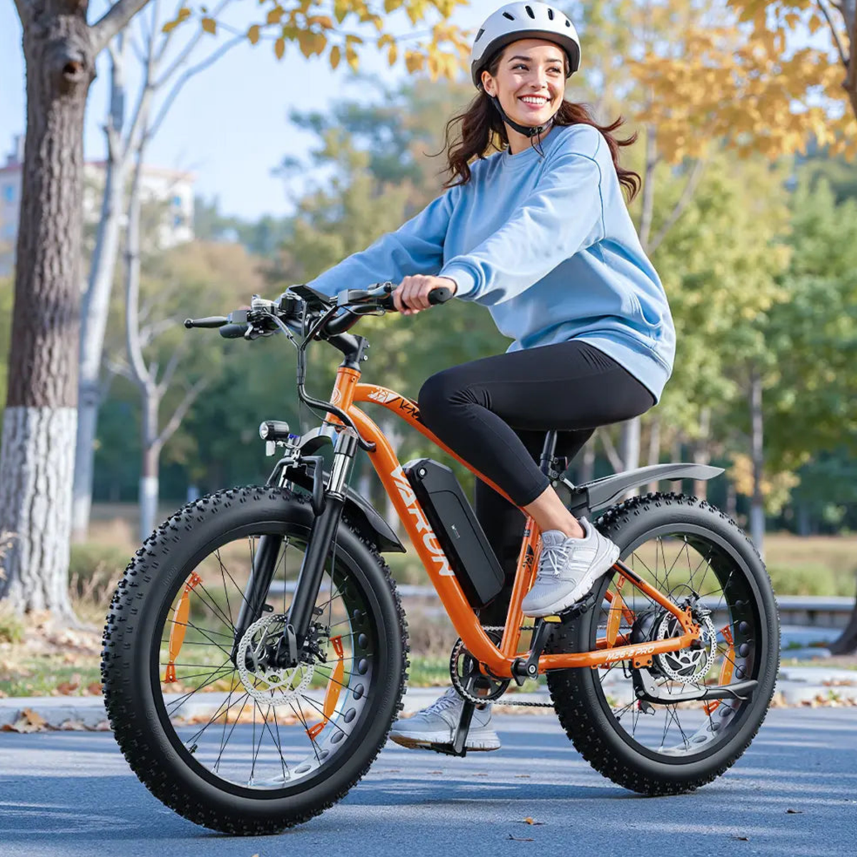 Donna alla guida della fat bike elettrica VARUN M26-2 Pro in colore arancione con il casco in un parco